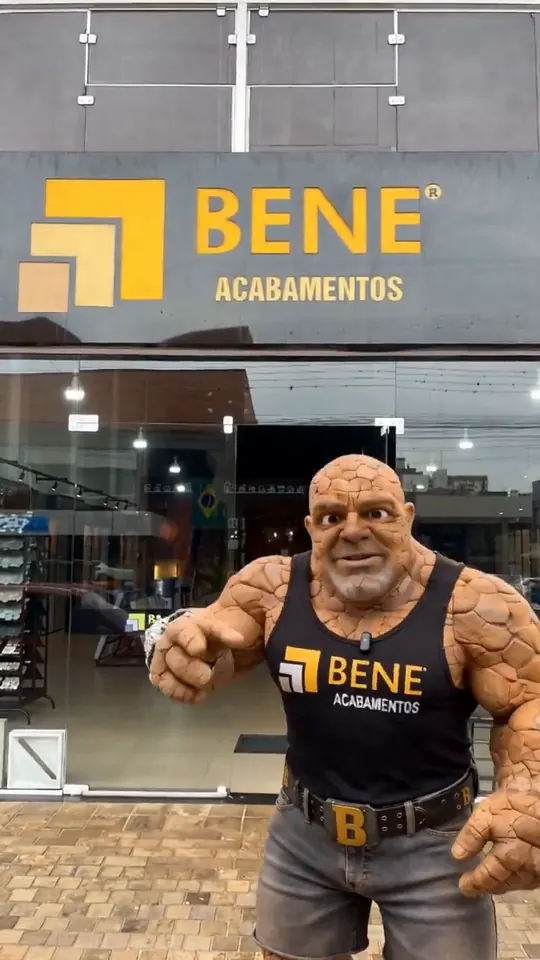 Personagem de pele de pedra com camiseta da Bene Acabamentos em frente à fachada da loja.